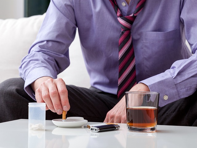 Man in pak zittend aan tafel met een glas alcohol, sigaretten en pillen