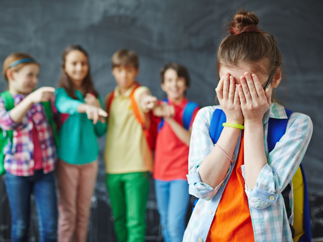 Meisje op de voorgrond met haar handen voor haar gezicht, op de achtergrond staan vier kinderen te lachen en wijzen